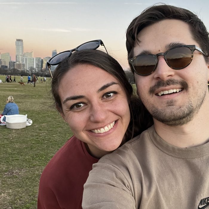 Andrea and Alex taking a selfie in a park at dusk, a city skyline behind them
